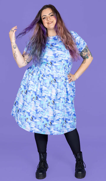 a model with purple hair wearing Winter Friends Stretch Belted Tea Dress with Pockets paired with black boots and tights. The jumpsuit is a light blue colour with ice burgs, seals, polar bears and penguins on. The model is smiling and flicking her hair in front of a purple background.