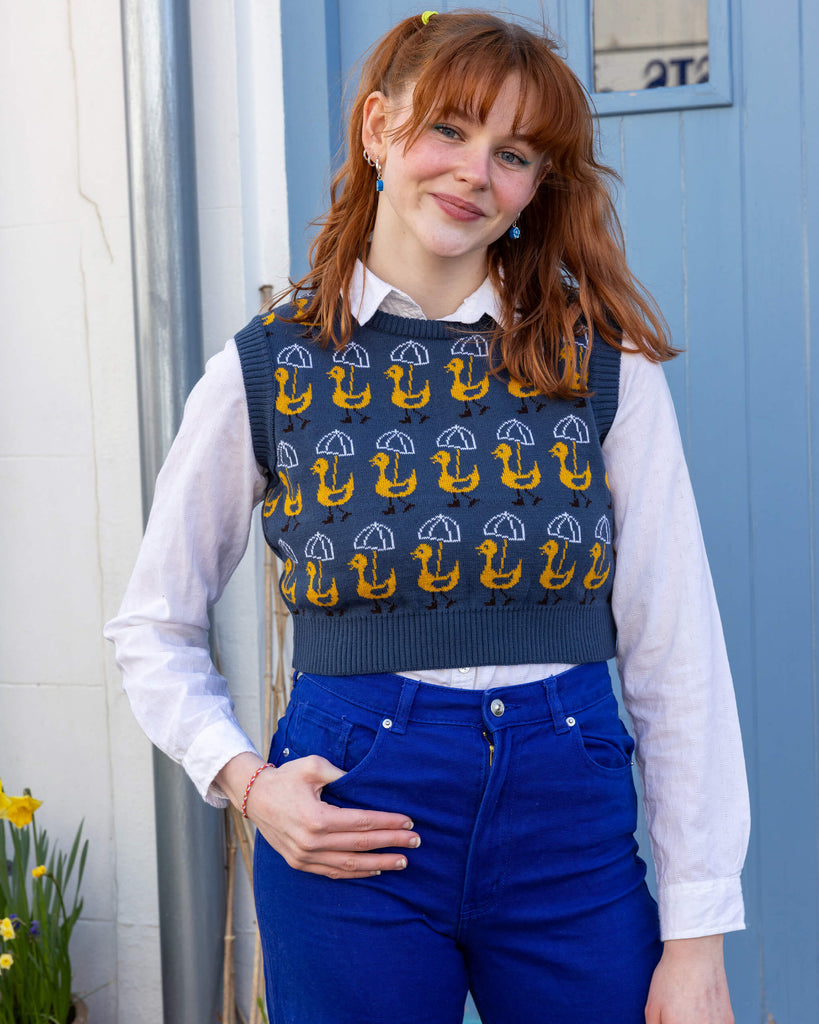 Model with red hair wearing Run & Fly Nice Weather for Ducks Cropped Knitted Tank Top paired with yellow hair accessories, blue earrings, white shirt and bright blue jeans. The tank top is a dusty dark blue colour with rows of yellow ducks holding white umbrellas printed on it. The tank top is a cropped style. Model is looking at camera and smiling