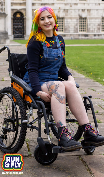 Rainbow haired femme person sat in a wheelchair smiling into camera wearing blue stretch denim dungaree shorts and a blue rainbow dinosaur jumper underneath.