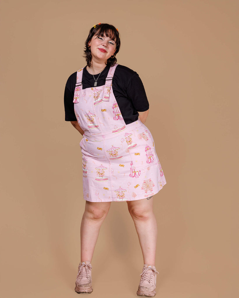 A model wears the Pink Fairground Stretch Twill Pinafore Dress by Run & Fly, a small ethical slow fashion brand. The light pink dress features playful fairground-themed illustrations of carousels, ferris wheels, and tickets. Paired with a black T-shirt, the outfit has a fun, nostalgic vibe. The model smiles with a relaxed stance, embodying the dress’s whimsical, retro feel.