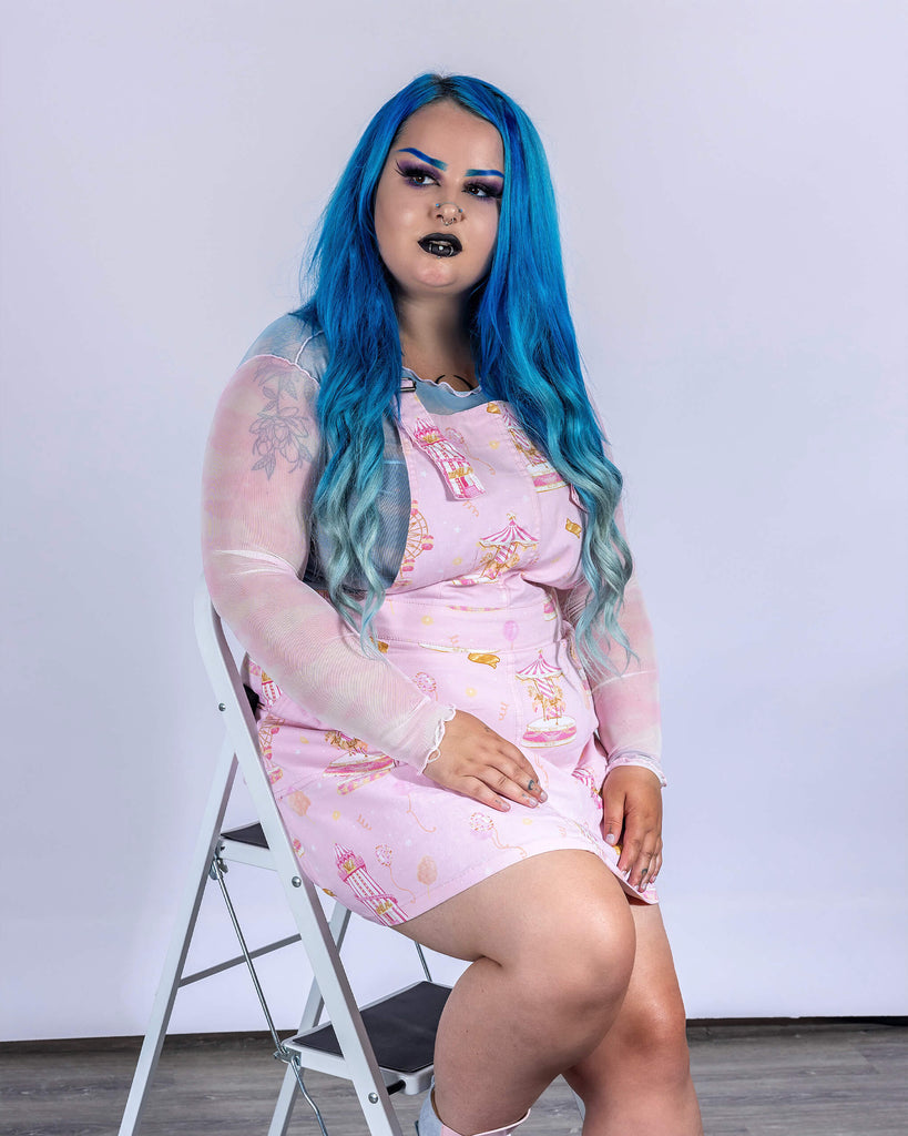 Model seated in Run & Fly’s Pink Fairground Stretch Twill Pinafore Dress. The whimsical fairground print, featuring carousels and ticket motifs, pops against the pastel pink fabric. This ethical, slow fashion piece adds a nostalgic, playful vibe, perfect for alternative and retro-inspired styles.