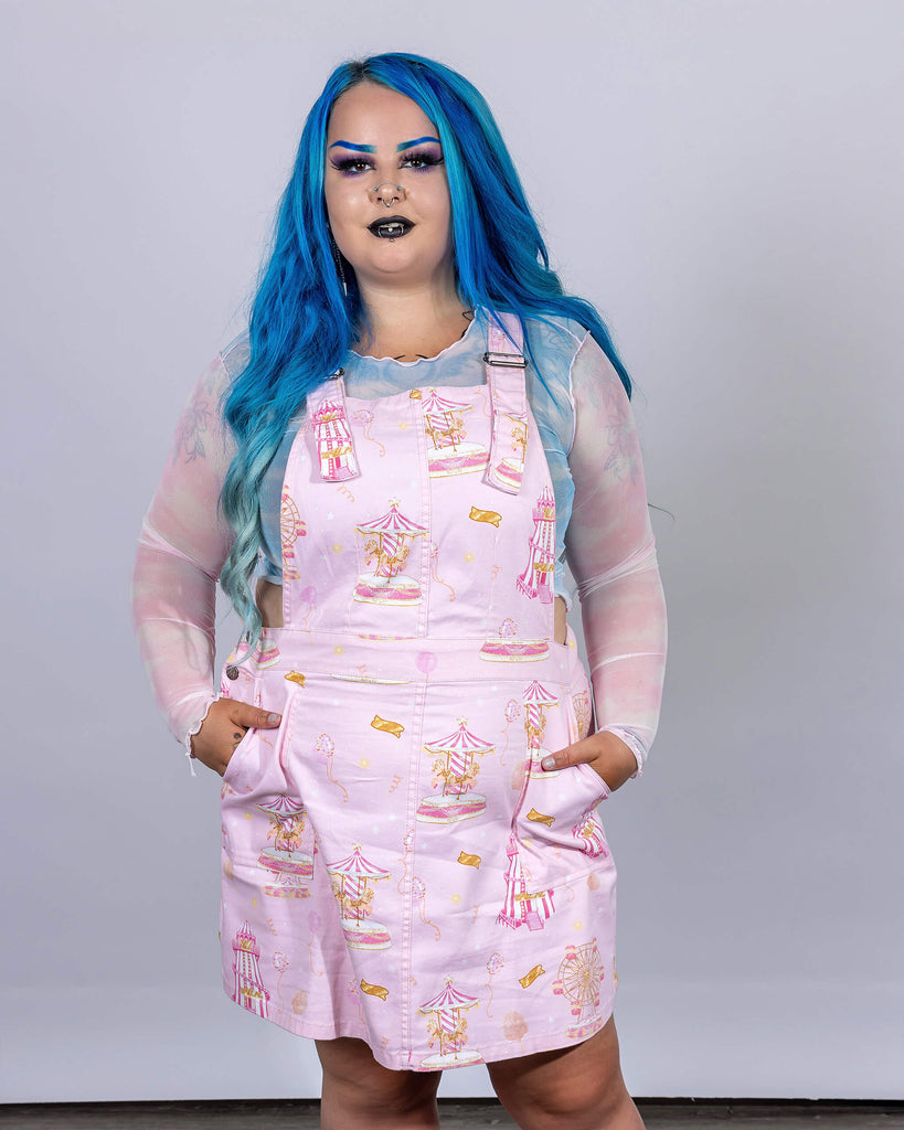 Model poses in Run & Fly’s Pink Fairground Stretch Twill Pinafore Dress, decorated with nostalgic fairground illustrations like carousels and balloons. The light pink dress captures a playful spirit, designed by this ethical, slow fashion brand to bring a fun, vintage vibe to modern wardrobes.
