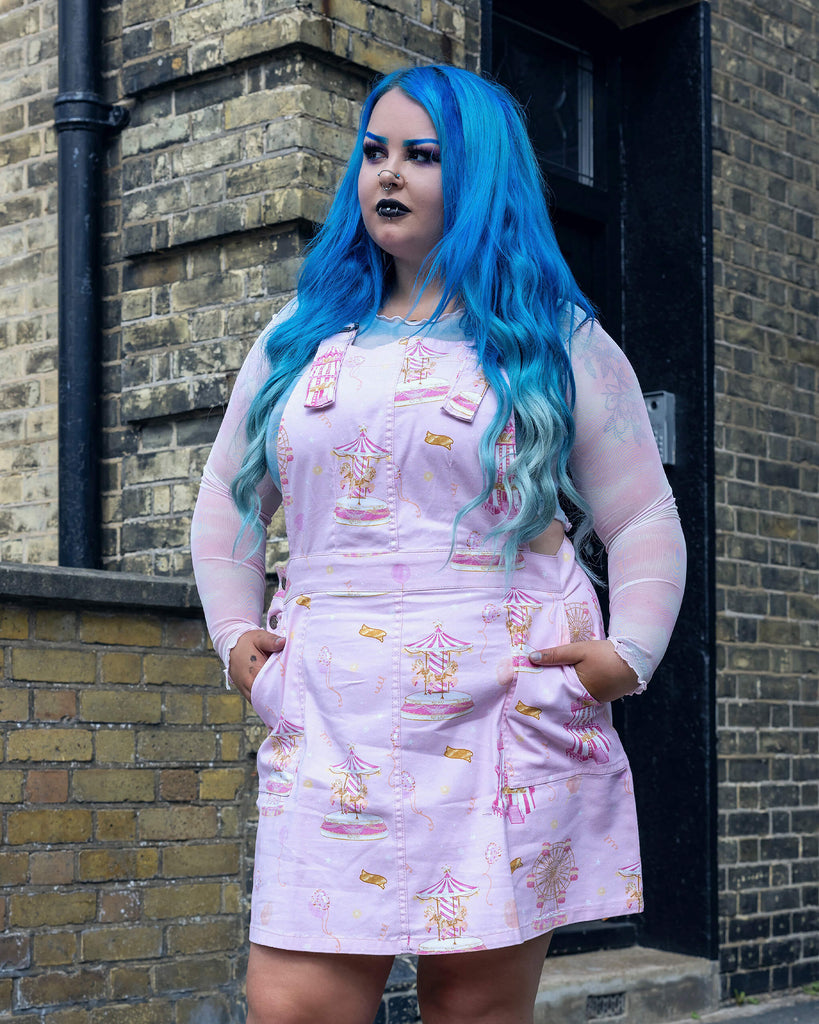  Full view of the Pink Fairground Stretch Twill Pinafore Dress by Run & Fly, featuring a whimsical fairground print. The model with vibrant blue hair and gothic makeup contrasts beautifully with the soft pink, showcasing the dress's playful, retro feel. Run & Fly embodies ethical, slow fashion with a unique twist.