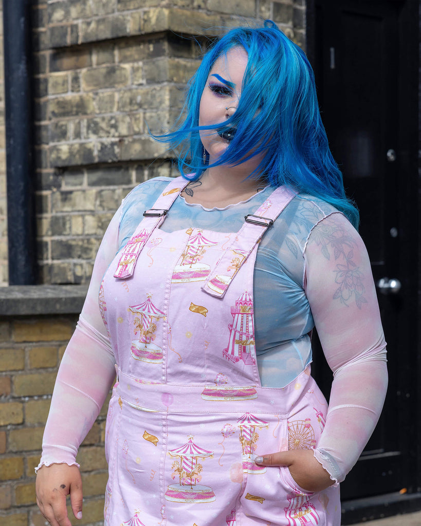 Model with blue hair wears Run & Fly’s Pink Fairground Stretch Twill Pinafore Dress. The fairground-themed print of carousels and tickets stands out on the pastel fabric. Styled with a sheer top, this ethical, slow fashion piece exudes a playful, vintage-inspired charm that’s perfect for alternative fashion lovers.