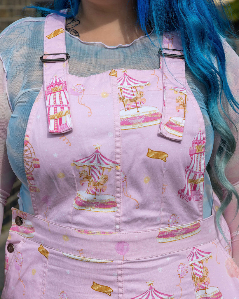  Close-up of Run & Fly’s Pink Fairground Stretch Twill Pinafore Dress, highlighting the playful fairground print with carousels, balloons, and tickets on a soft pink background. The print adds a whimsical, retro touch to this ethical slow fashion piece, designed for fans of unique, nostalgic styles.