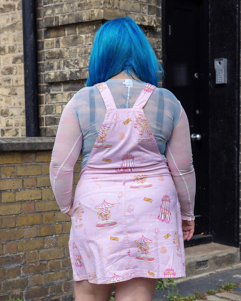 Back view of the Pink Fairground Stretch Twill Pinafore Dress by Run & Fly, displaying the dress’s playful fairground-themed print, including carousels and ferris wheels. The model’s blue hair contrasts with the pastel dress, showing off the brand’s fun, ethical style. This slow fashion piece captures vintage amusement park charm in a modern silhouette.