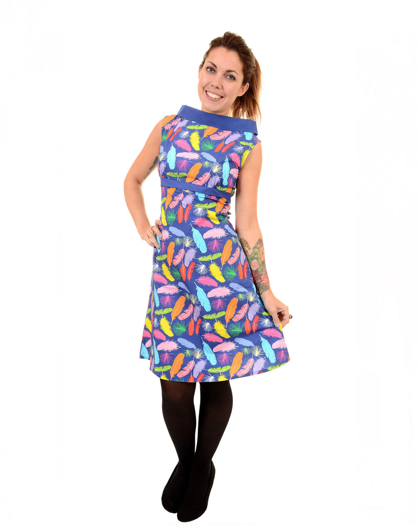 Model is stood in front of a white studio background wearing the feather 60's dress with tights underneath and black heels. They are facing the camera and posing with one hand on their hip and the other holding out the bottom of the dress. They are looking into the camera and smiling.