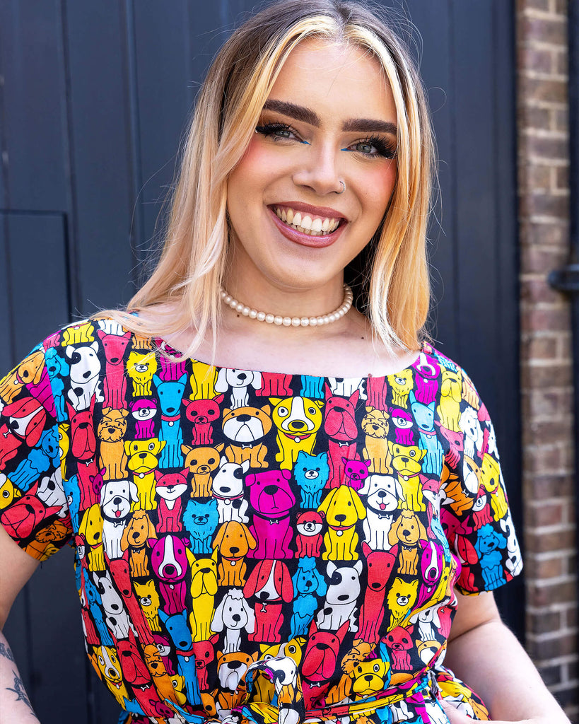Close up of model with long blonde hair wearing Finding Fox Dog Tea Stretch Dress with Pockets paired with a pearl choker. The dress has a black background with various dog breeds in bright yellow, blue, pink, orange, red and white printed all over. Model is smiling at the camera with thumbs in pockets.