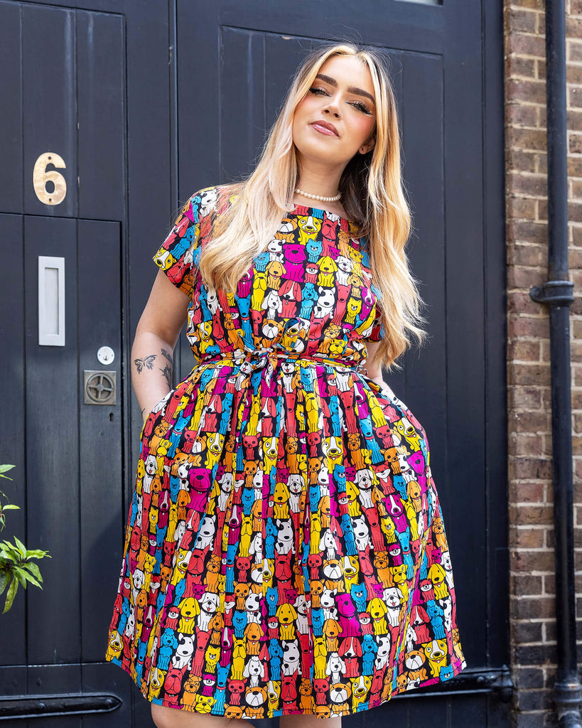 Model with long blonde hair wearing Finding Fox Dog Tea Stretch Dress with Pockets paired with a pearl choker and black trainers with white socks. The dress has a black background with various dog breeds in bright yellow, blue, pink, orange, red and white printed all over. Model is posing with hands in pockets.
