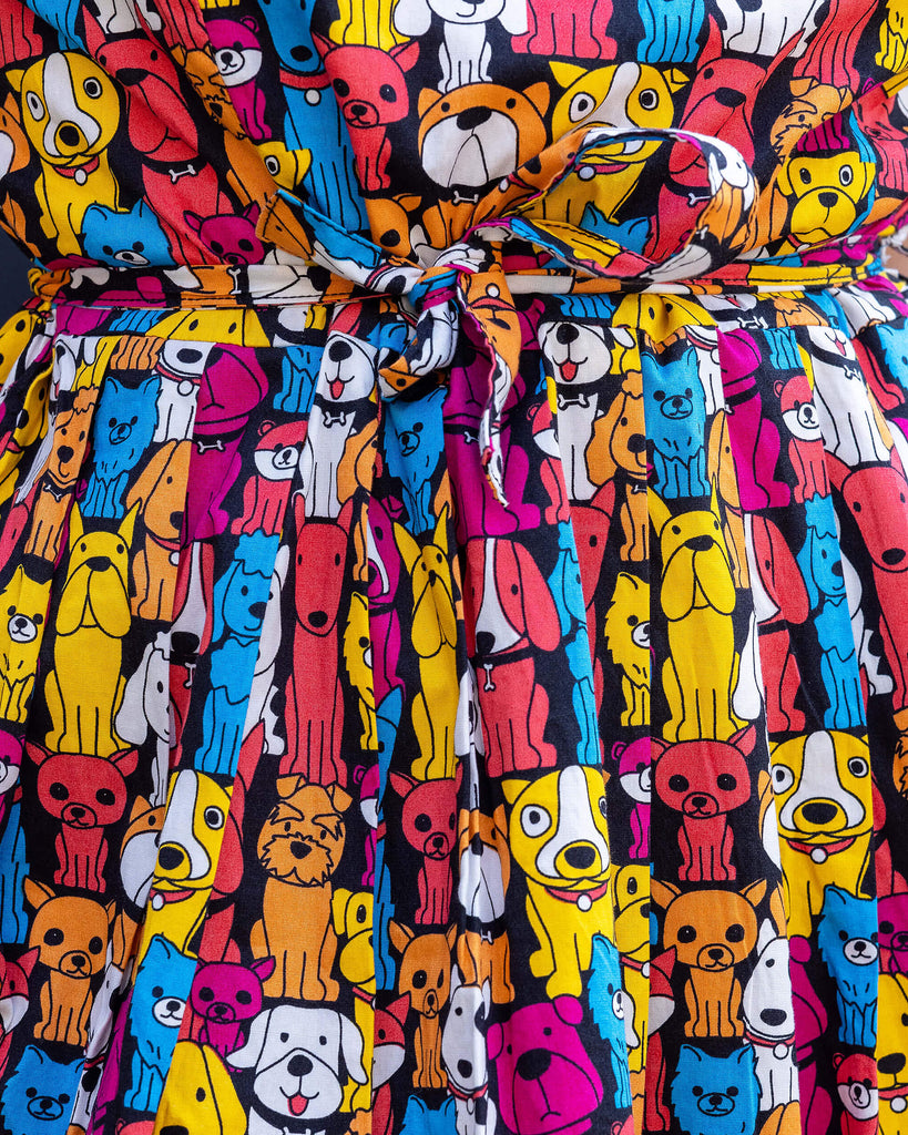 Close up of model wearing Finding Fox Dog Tea Stretch Dress with Pockets. The dress has a black background with various dog breeds in bright yellow, blue, pink, orange, red and white printed all over. Model has back to camera to show matching fabric belt tied in a bow around waist.