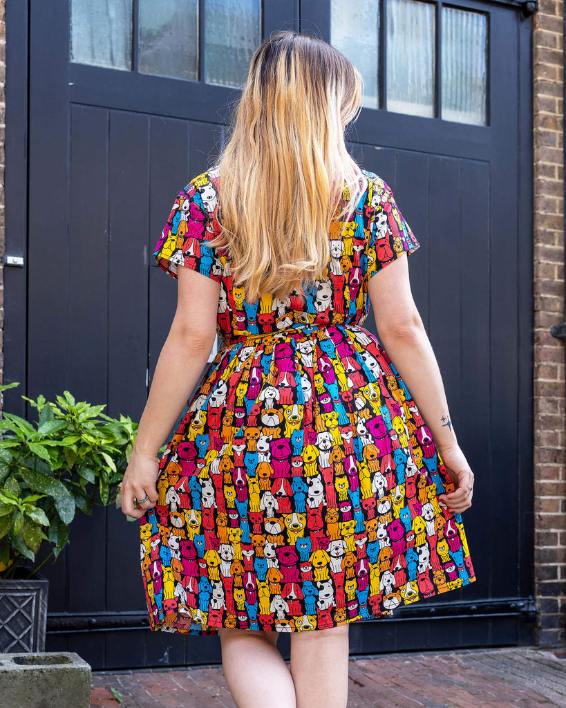 Close up of model wearing Finding Fox Dog Tea Stretch Dress with Pockets. The dress has a black background with various dog breeds in bright yellow, blue, pink, orange, red and white printed all over. Model has back to camera to show matching fabric belt tied in a bow around waist.