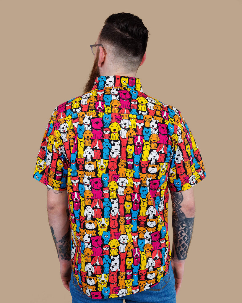 Declan a white bearded tattooed man in his 20's with brown hair and glasses is smiling at camera wearing a multicoloured shirt with dogs on and blue jeans on a beige backdrop shot in studio.