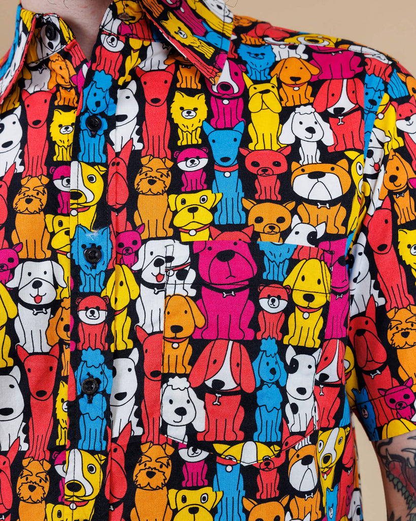 A close up of Declan a white bearded tattooed man in his 20's with brown hair and glasses is smiling at camera wearing a multicoloured shirt with dogs on and blue jeans on a beige backdrop shot in studio