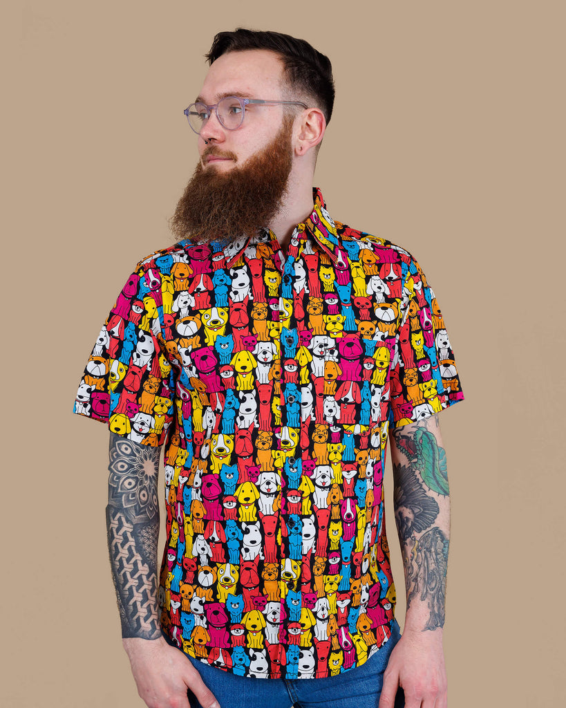 Declan a white bearded tattooed man in his 20's with brown hair and glasses is smiling at camera wearing a multicoloured shirt with dogs on and blue jeans on a beige backdrop shot in studio.