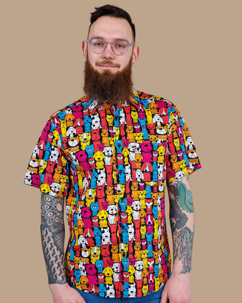 Declan a white bearded tattooed man in his 20's with brown hair and glasses is smiling at camera wearing a multicoloured shirt with dogs on and blue jeans on a beige backdrop shot in studio.