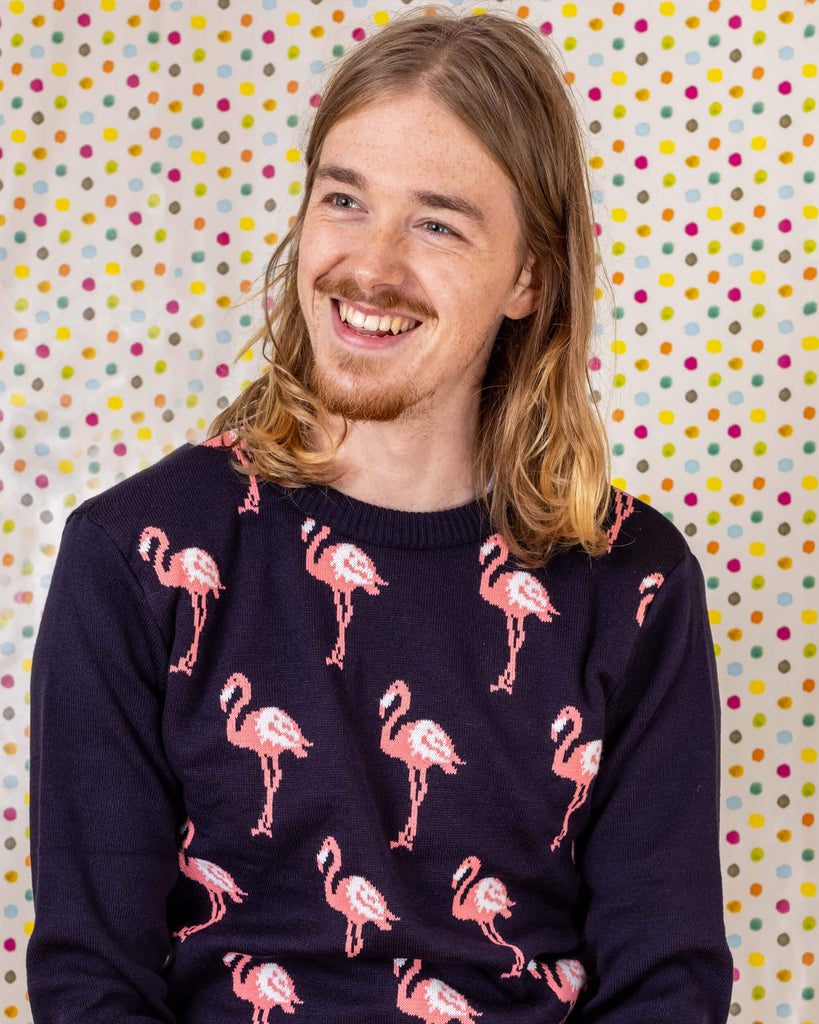 Jack is sat down in front of a multicoloured dotted background wearing the navy blue flamingo Jumper with jeans. He is facing the camera but looking off to the left and smiling.