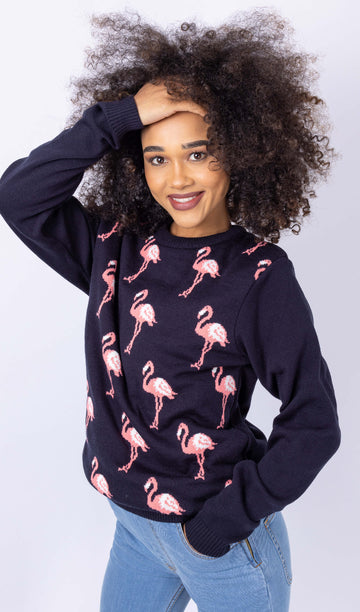 Model is stood in front of a white studio background wearing the flamingo jumper and light wash denim jeans. They are facing the left with one hand on their head and the other in their front pocket, they are looking directly into the camera and smiling. Photo is cropped from the thighs up.