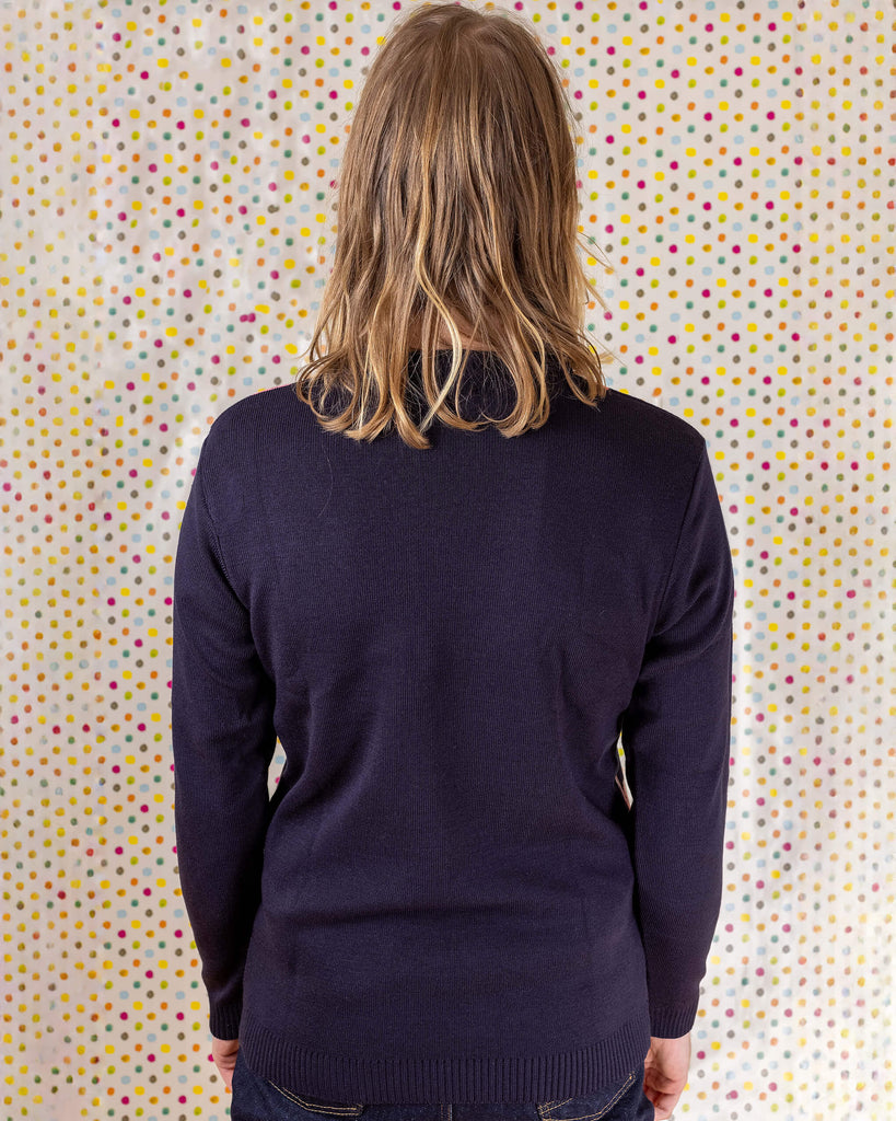 Model is stood in front of a multicoloured dotted background wearing the flamingo jumper with blue jeans. They are facing away from the camera with both hands resting by their sides. Photo is cropped from the thighs up.