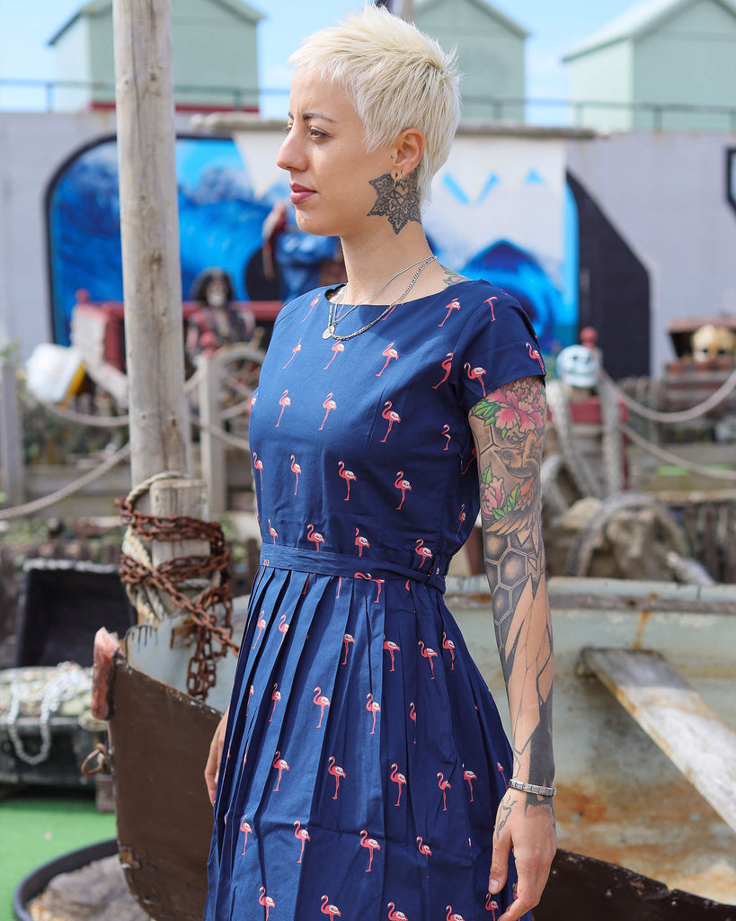 Model is stood outside in a seaside park area wearing the flamingo tea party dress. They are facing to the left with both hands resting by their sides. Photo is cropped from the knees up.