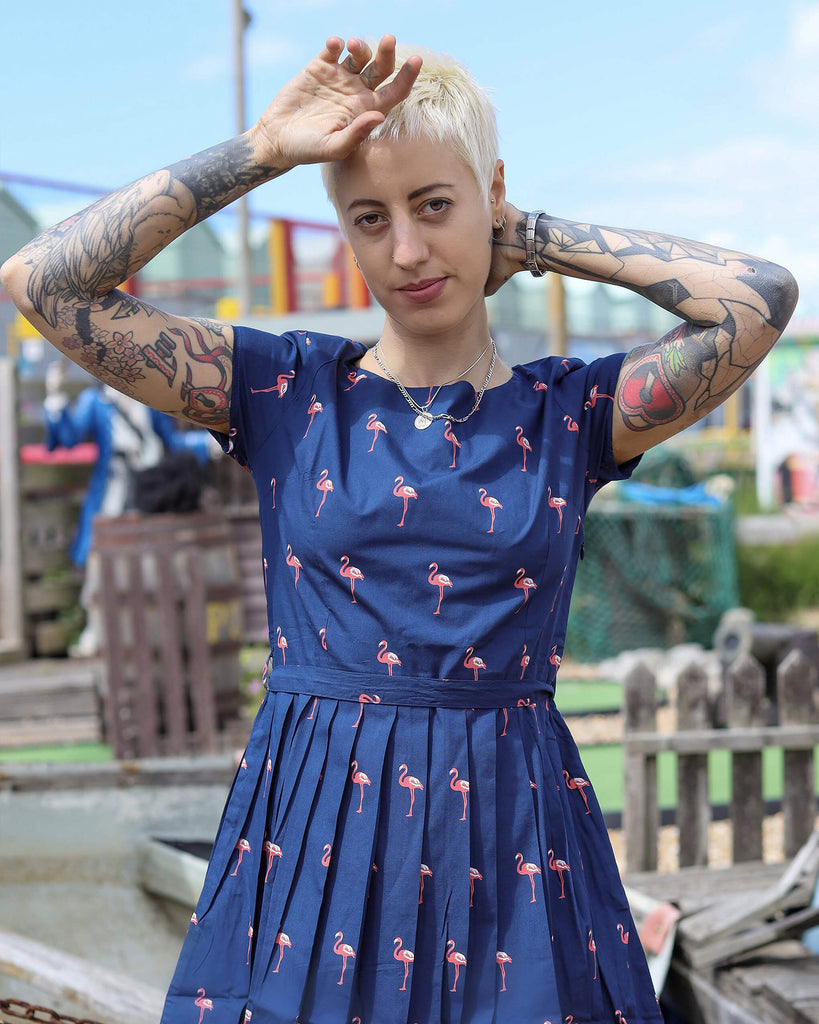 Model is stood outside in a seaside park area wearing the flamingo tea party dress. They are facing the camera posing with one hand behind their head and the other in front of their head. Photo is cropped from the knees up.