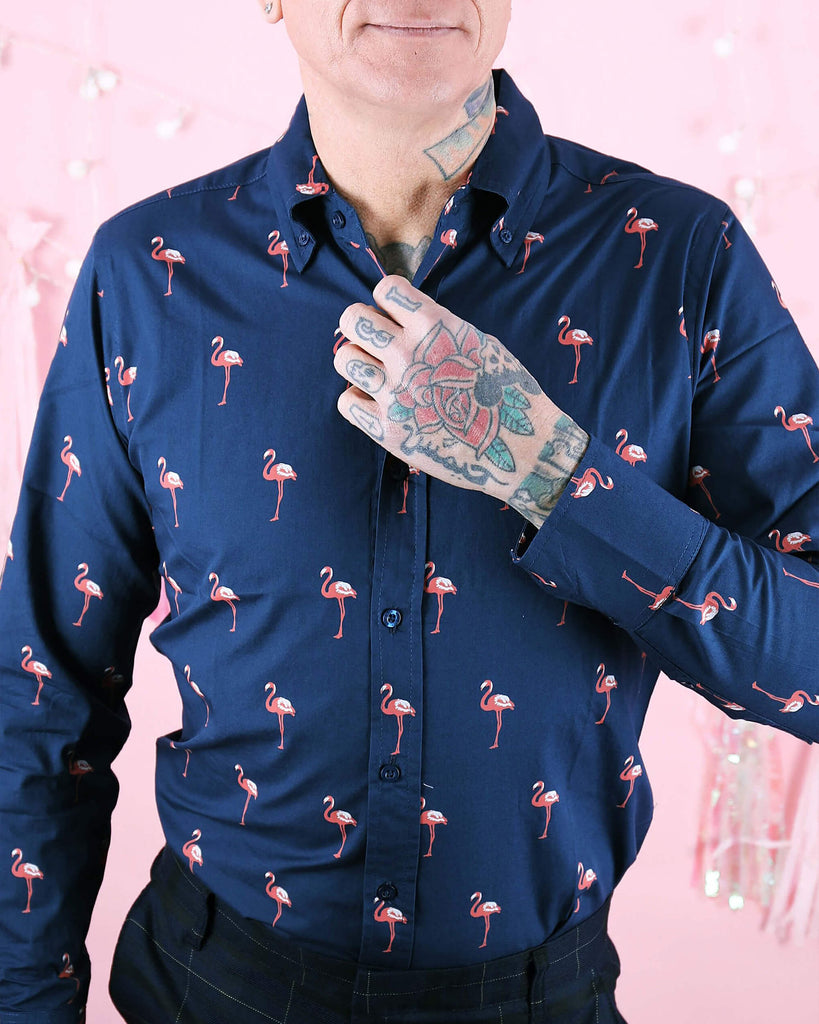 Close-up of a tattooed model wearing Run & Fly’s Flamingo Print Long Sleeve Shirt in navy blue, featuring small pink flamingos printed all over. The model adjusts the shirt collar, showing the button-down detail and vibrant design. Perfect for quirky, statement fashion. Run & Fly is a small, ethical, slow fashion brand known for unique, fun prints.