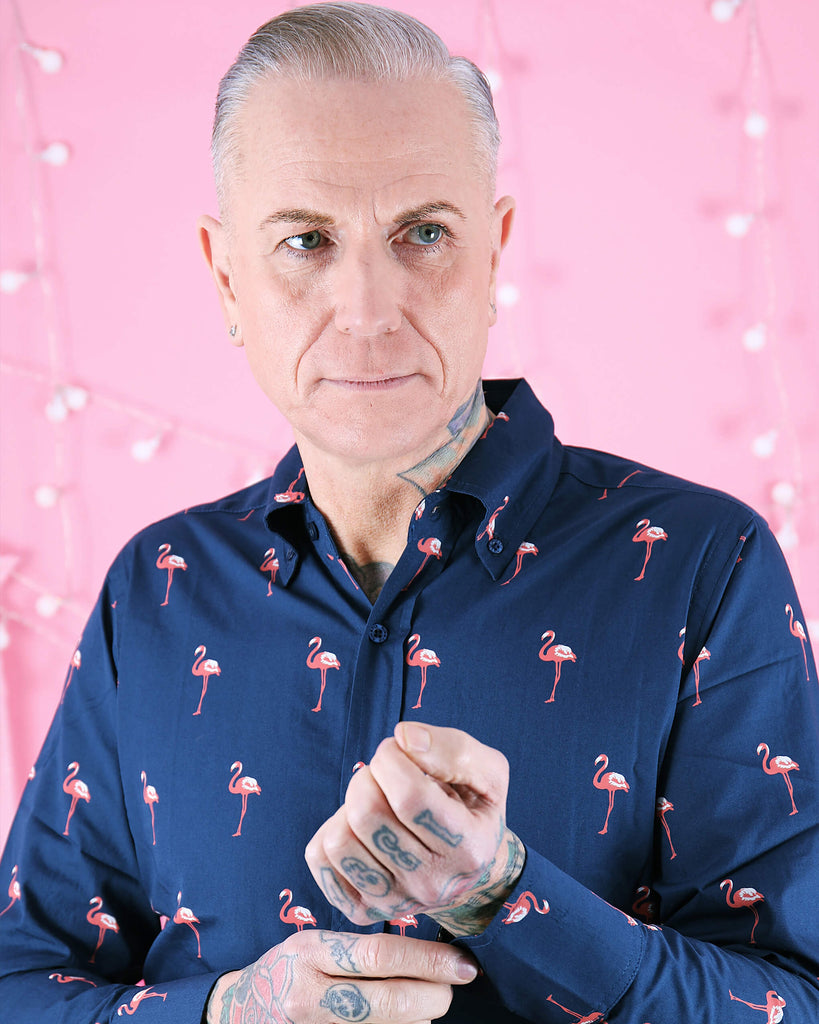 Portrait of a model wearing Run & Fly’s Flamingo Print Long Sleeve Shirt, featuring a bold flamingo print on navy. The model has tattoos and adjusts the cuff, highlighting the shirt’s tailored fit. This unique piece by ethical slow fashion brand Run & Fly adds a playful twist to classic menswear.