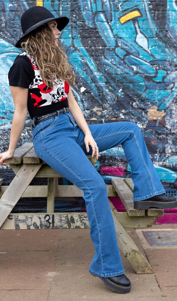 Hannah is on a picnic bench in front of a graffiti wall wearing the unisex stonewashed bellbottom flares leg high waisted jeans with the Seeing Spots Dalmatian Knitted Tank Top, black boots and a black hat. They are facing to the right posing leaning back on one hand and with one leg up, they are looking to the right.
