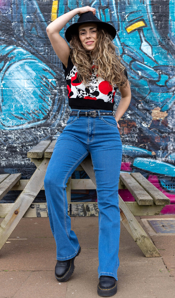 Hannah is leaning against a picnic bench in front of a graffiti wall wearing the unisex stonewashed bellbottom flares leg high waisted jeans with the Seeing Spots Dalmatian Knitted Tank Top, black boots and a black hat. They are facing the camera posing with one hand up on their hat whilst the other resting on the bench behind behind them whilst smiling.