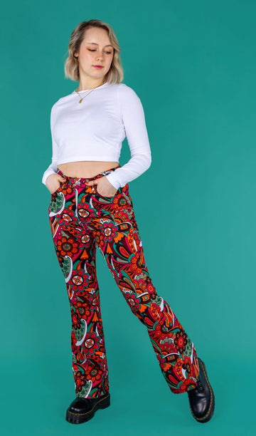 Amy a blonde model with bobbed hair is wearing a white long sleeved cropped tee with jazzy 70's swirly flares with black boots on a green background 