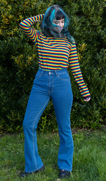 Faeryn is stood in a garden area wearing the unisex stonewashed bellbottom flares leg high waisted jeans with the Retro Rainbow Brights Repeat Striped Long Sleeved T Shirt and black trainers. They are facing forward posing with one leg bent out to show off the flares and one hand behind their head whilst looking down to the right.