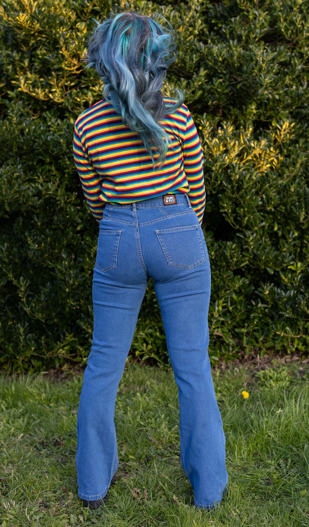 Faeryn is stood in a garden area wearing the unisex stonewashed bellbottom flares leg high waisted jeans with the Retro Rainbow Brights Repeat Striped Long Sleeved T Shirt and black trainers. They are facing away from the camera with both hands in front of them.
