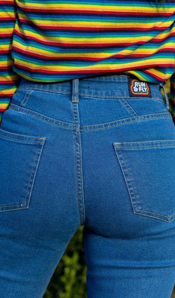 Close up shot of the back stitching and run and fly waist label on the unisex stonewashed bellbottom flares leg high waisted jeans paired with the retro rainbow brights repeat striped long sleeved tshirt.