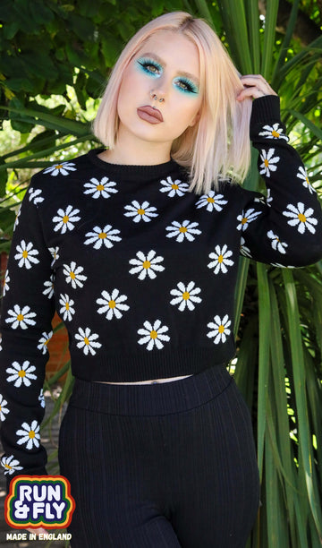 Model is stood in a green garden area wearing the daisy chain black cropped jumper with black trousers. They are facing the camera with one hand in their hair and the other by their side. Photo is cropped from the hips up.