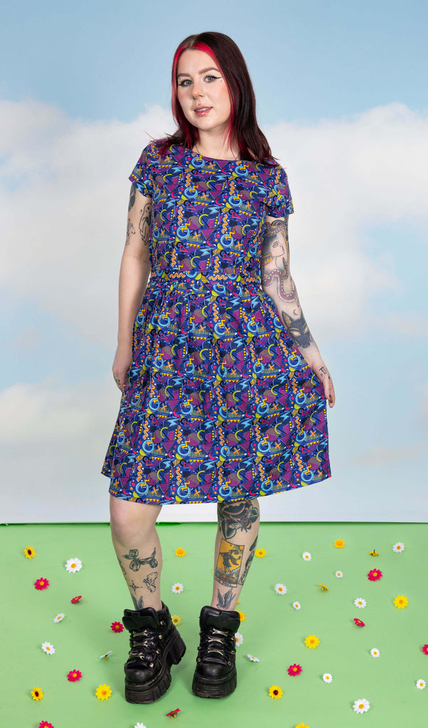 Florence is stood in a photography studio in Hove in front of blue sky backdrop on a green floor with flowers scattered wearing 90's Arcade Stretch Belted Tea Dress with Pockets with chunky black shoes. The print features classic 90's style shapes, squiggles and doodles in green, orange, pink, purple and light blue all on a dark blue background. Florence is posing and smiling at the camera with one leg slightly bent.