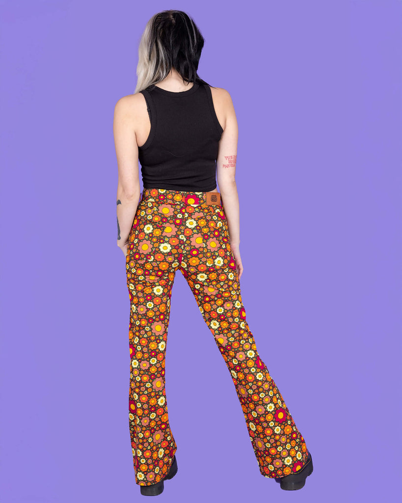 Model is wearing Ditsy Floral Bell Bottom Flares paired with a black crop top and black boots. The flares are a dark green and are covered in red, orange and yellow flowers. The model is posing with their back to the camera and the background of the photo is purple.