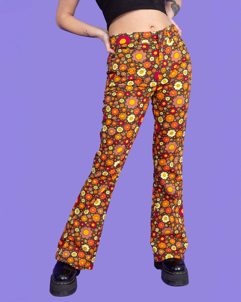 Model is wearing Ditsy Floral Bell Bottom Flares paired with a black crop top and black boots. The flares are a dark green colour and covered in red, orange and yellow flowers. The model is posing in front of a purple background facing the camera.