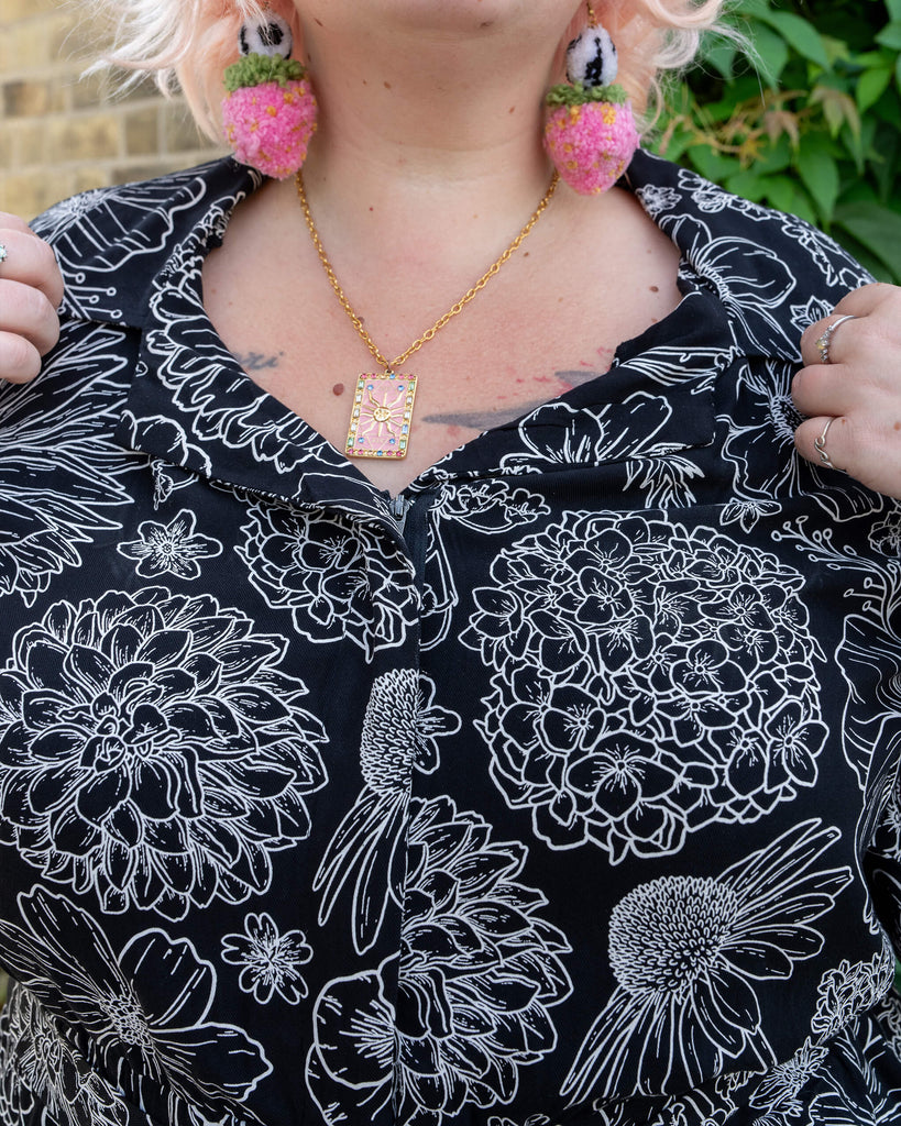 Close-up of the front of a black and white floral jumpsuit by Run & Fly. The model, with pastel pink hair and strawberry earrings, wears a delicate gold necklace. The intricate floral designs in white stand out against the black background of the jumpsuit. Perfect for a slow fashion wardrobe, the rayon fabric is lightweight and breathable.