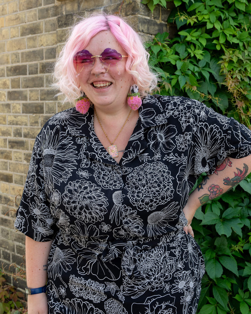 A model with pastel pink hair smiles brightly while wearing a black and white floral jumpsuit by Run & Fly. The bold floral design stands out against the black background of the rayon fabric. The model pairs the outfit with heart-shaped sunglasses, strawberry earrings, and a necklace, creating a playful and vibrant look. The background features a brick wall and lush green leaves, emphasising the ethical slow fashion brand’s charm.