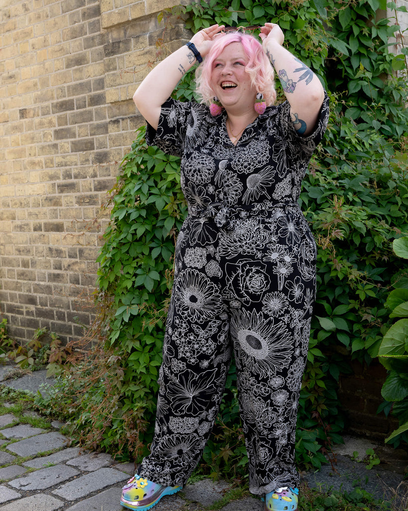 A model with pastel pink hair laughs and adjusts their heart-shaped sunglasses while wearing a black and white floral jumpsuit by Run & Fly. The jumpsuit features an intricate floral design on lightweight rayon fabric. They pair it with rainbow platform shoes for a quirky, colourful touch. The relaxed, comfortable fit of the jumpsuit is ideal for alternative and slow fashion lovers. The leafy and brick background complements the playful energy of the scene.
