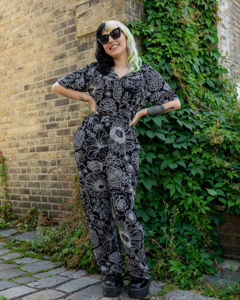 A model with black and lime hair stands confidently, wearing a black and white floral jumpsuit by Run & Fly. The detailed white floral design contrasts beautifully with the black rayon fabric. The model completes the look with black platform boots and oversized sunglasses. The casual yet bold look is perfect for alternative and slow fashion enthusiasts. The background features a brick wall and leafy greenery, adding a natural touch to the shoot.