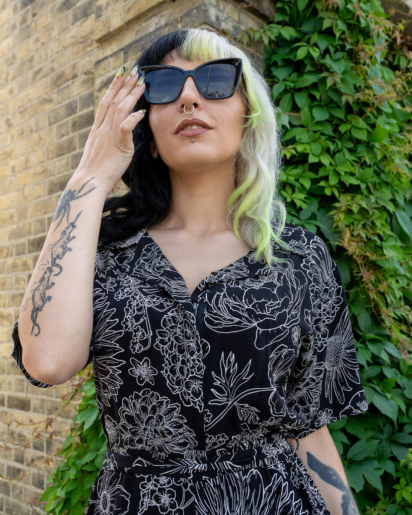 A model with black and lime green hair adjusts their oversized sunglasses while wearing a black and white floral jumpsuit by Run & Fly. The jumpsuit features a bold floral design in white on a black background. The model’s tattooed arm and confident pose add a stylish, alternative vibe to the scene. The background includes a brick wall and leafy greenery, highlighting the jumpsuit's ethical slow fashion appeal.