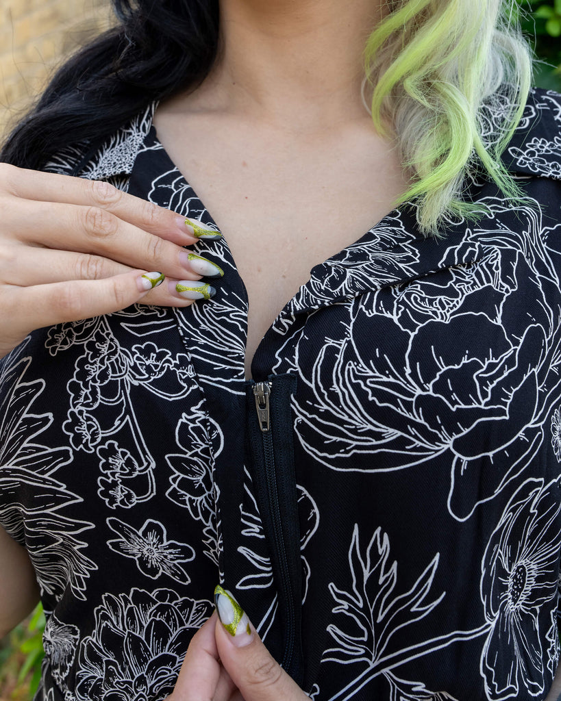Close-up of a model's hands adjusting the zipper on the front of a black jumpsuit by Run & Fly. The jumpsuit features a white floral pattern on a black background, crafted from lightweight rayon fabric. The model’s long, lime green nails and black and lime hair enhance the alternative fashion aesthetic. The detailed white floral design on the jumpsuit is perfect for those seeking ethical slow fashion.