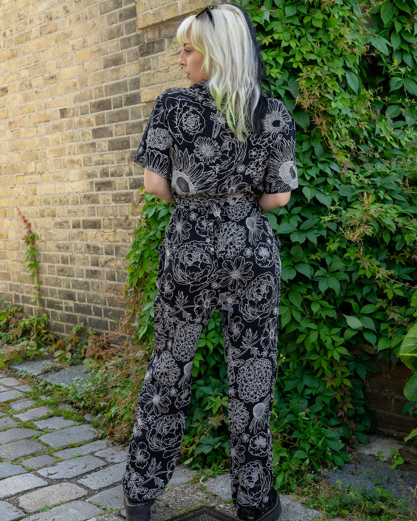 A model with black and lime green hair stands with their back turned, showing off the back of the black and white floral jumpsuit by Run & Fly. The detailed white floral designs on the black fabric create a striking contrast. The relaxed fit and cinched waist add a flattering silhouette. The leafy green background and brick wall complete the scene, showcasing the ethical slow fashion brand.