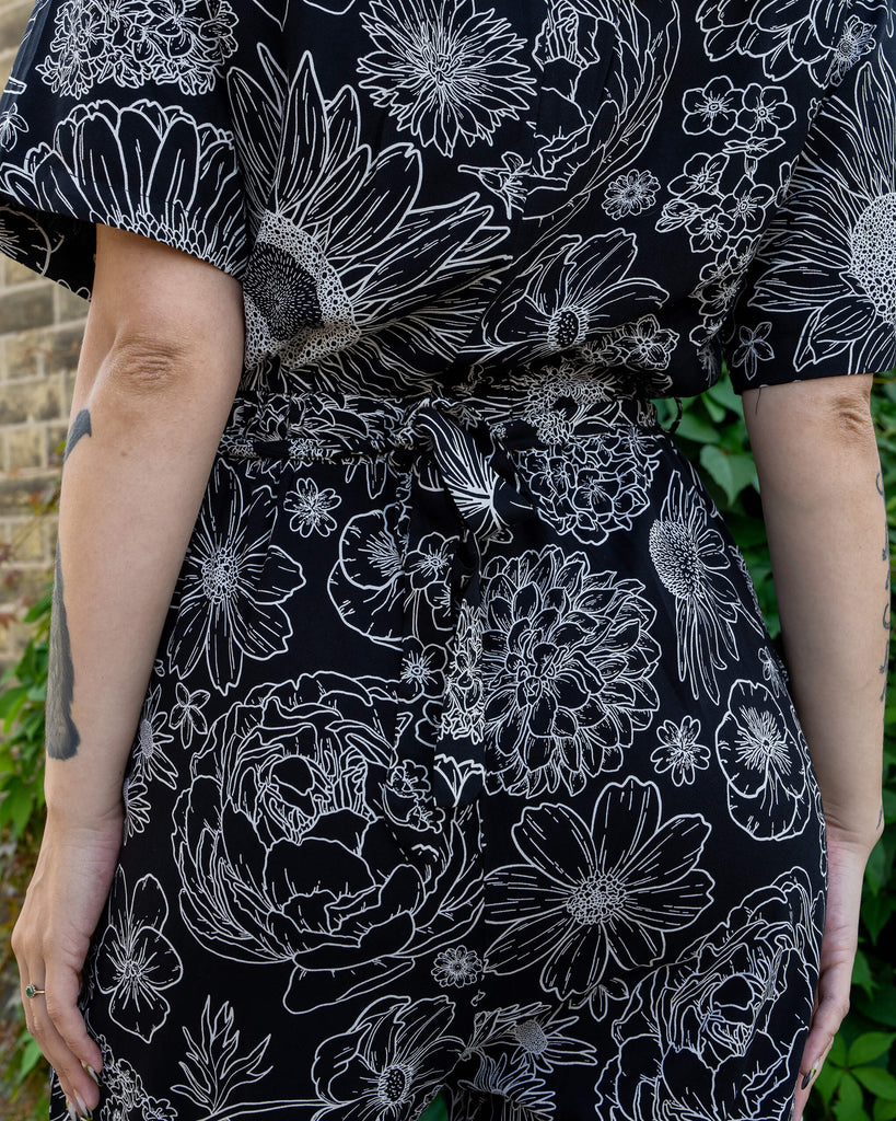 Close-up of a black jumpsuit by Run & Fly featuring a detailed white floral print. The image highlights the back of the garment with the waist tie and intricate flower designs. The jumpsuit is made from lightweight, ethical rayon fabric, perfect for slow fashion lovers. Model's arm tattoos and green leafy background add character.