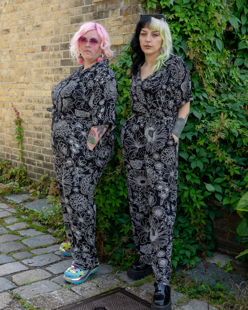 Two models pose against a brick wall in matching black and white floral jumpsuits by Run & Fly. The jumpsuits feature bold, detailed white flower illustrations. One model has pastel pink hair and rainbow platform shoes, while the other has black and lime hair with chunky black boots. Ethical slow fashion for alternative and floral fashion lovers.