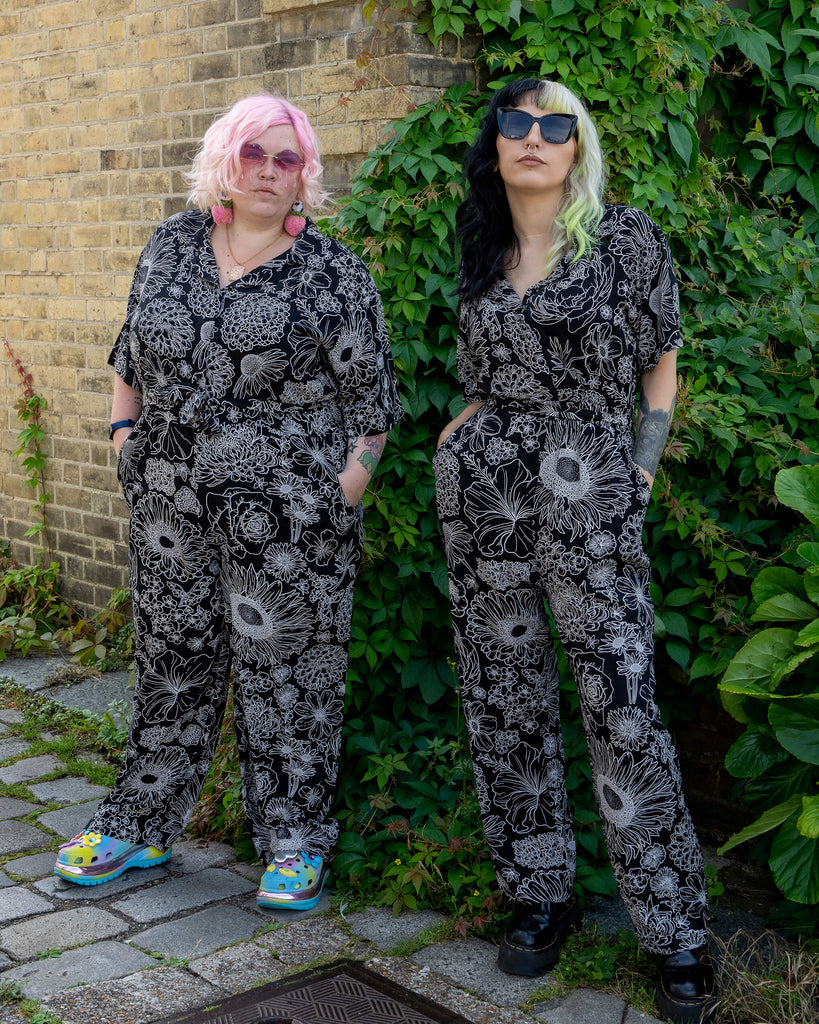 Two models pose in matching black and white floral jumpsuits by Run & Fly. The jumpsuits feature intricate white floral designs on lightweight rayon fabric. One model has pastel pink hair and wears colourful platform shoes, while the other has black and lime hair with black platform boots. The background of leafy greenery and a brick wall enhances the alternative and ethical slow fashion aesthetic.