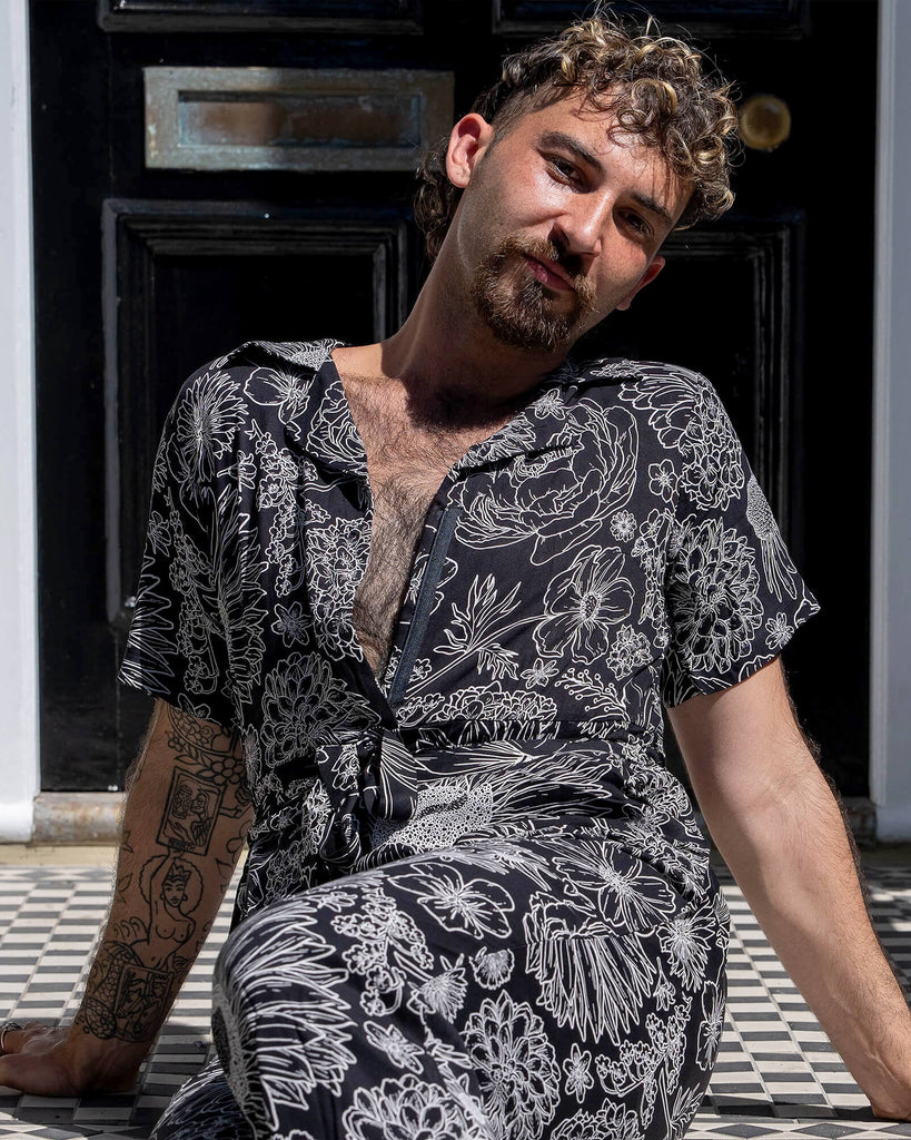 The Black and White Floral Rayon Jumpsuit worn by a masc non binary person with a brown curly mullet haircut and white trainers. They are sat in front of a doorway entrance with white ornate detailing, a black door, black ornate railing and black and white tiled steps. They are facing forward smiling leaning back on the steps. The black base jumpsuit features white floral outlines all over.