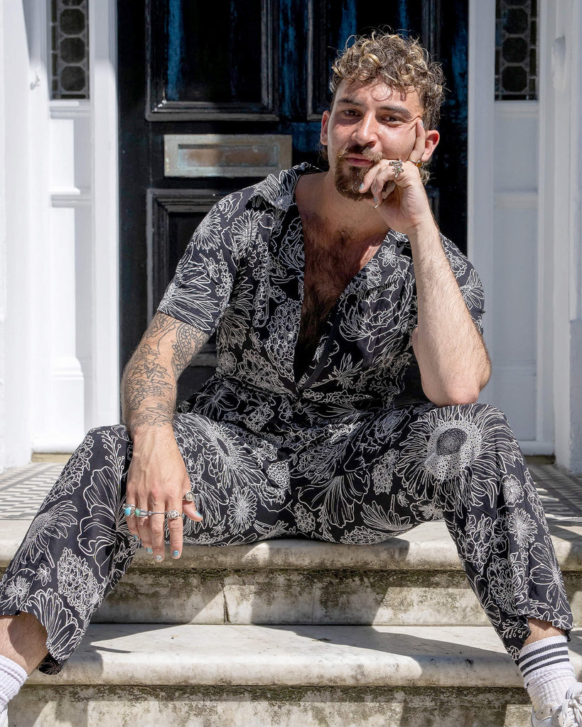 The Black and White Floral Rayon Jumpsuit worn by a masc non binary person with a brown curly mullet haircut and white trainers. They are sat in front of a doorway entrance with white ornate detailing, a black door, black ornate railing and black and white tiled steps. They are facing forward leaning forward onto their knee. The black base jumpsuit features white floral outlines all over.