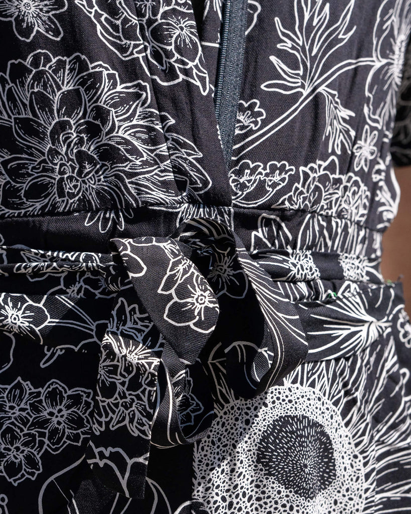 Close up of the Black and White Floral Rayon Jumpsuit zip up front and belt tie detailing worn by a masc non binary person. The black base jumpsuit features white floral outlines all over.