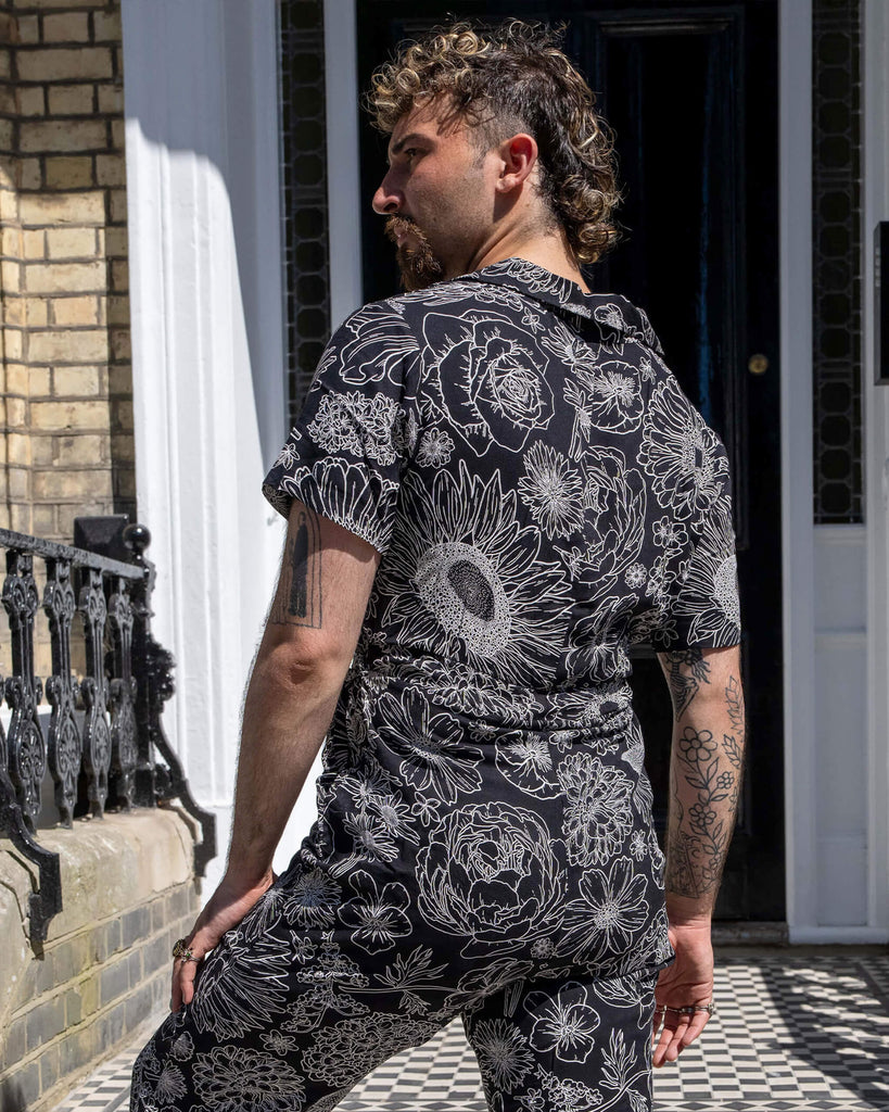The Black and White Floral Rayon Jumpsuit worn by a masc non binary person with a brown curly mullet haircut and white trainers. They are stood in front of a doorway entrance with white ornate detailing, a black door, black ornate railing and black and white tiled steps. They are facing away taking a step up the steps looking off to the left. The black base jumpsuit features white floral outlines all over.
